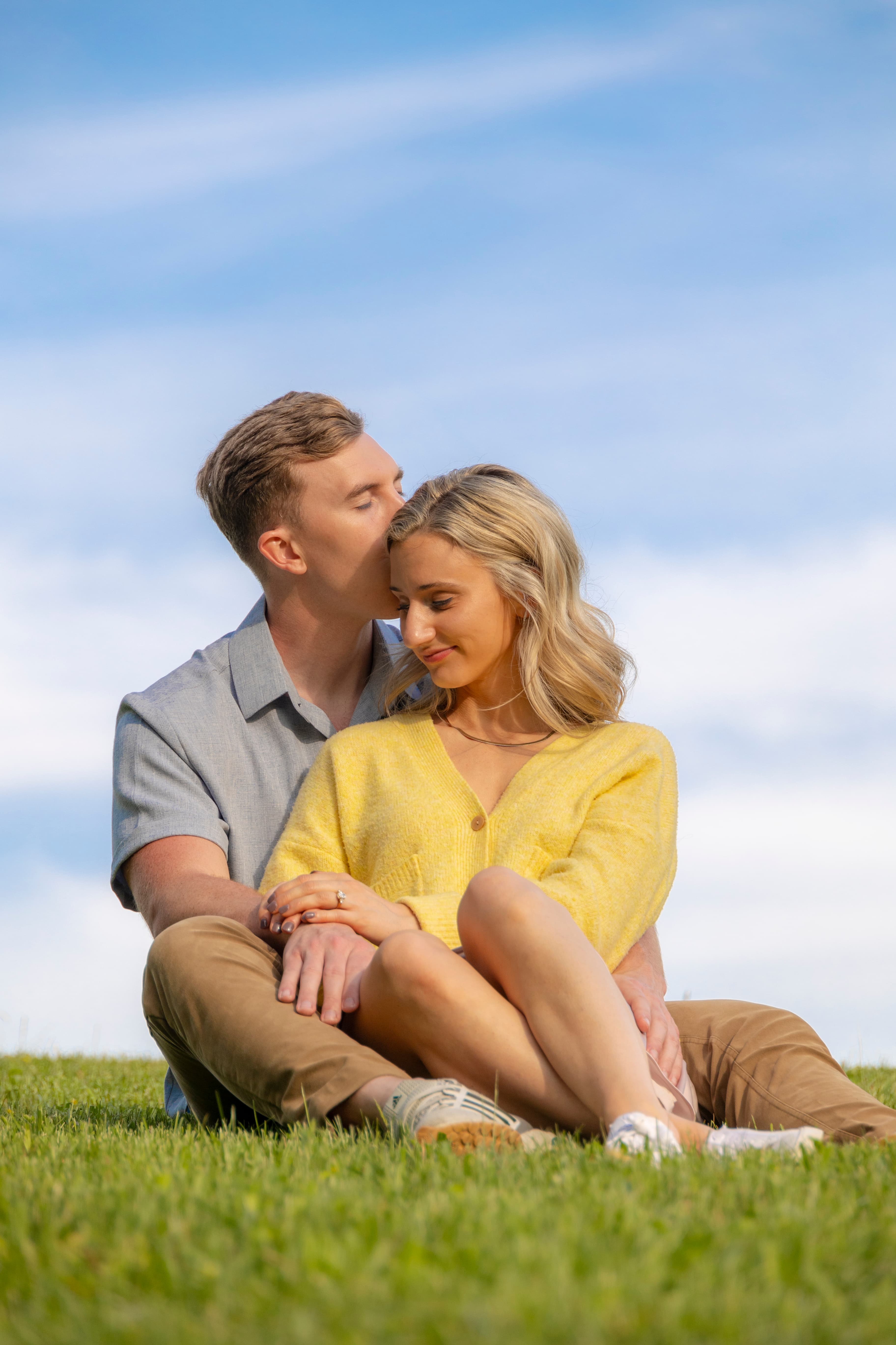 Couple sitting on grass, kissing her head, blue sky