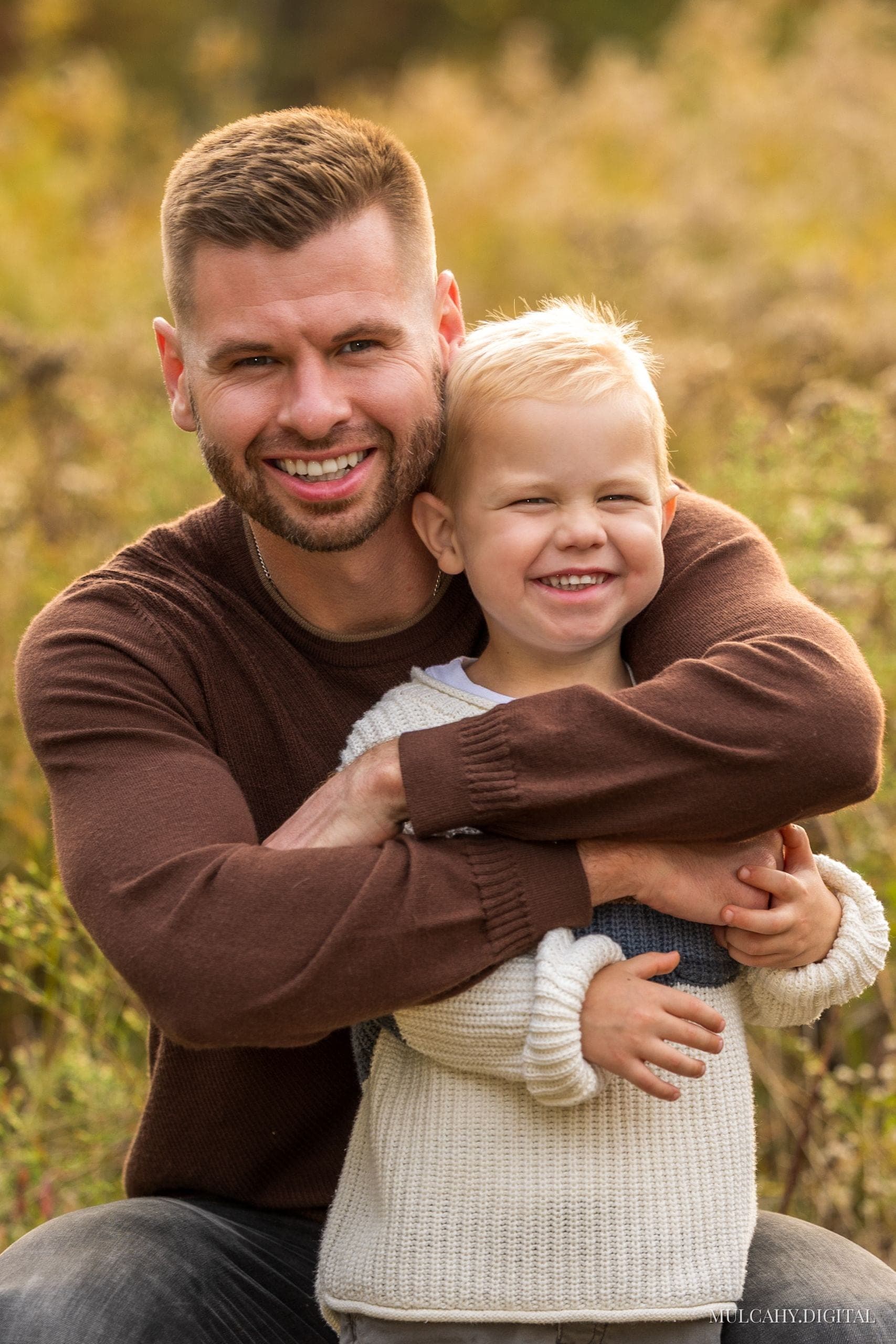Father in a brown sweater embracing his young son, both smiling in a golden autumn meadow