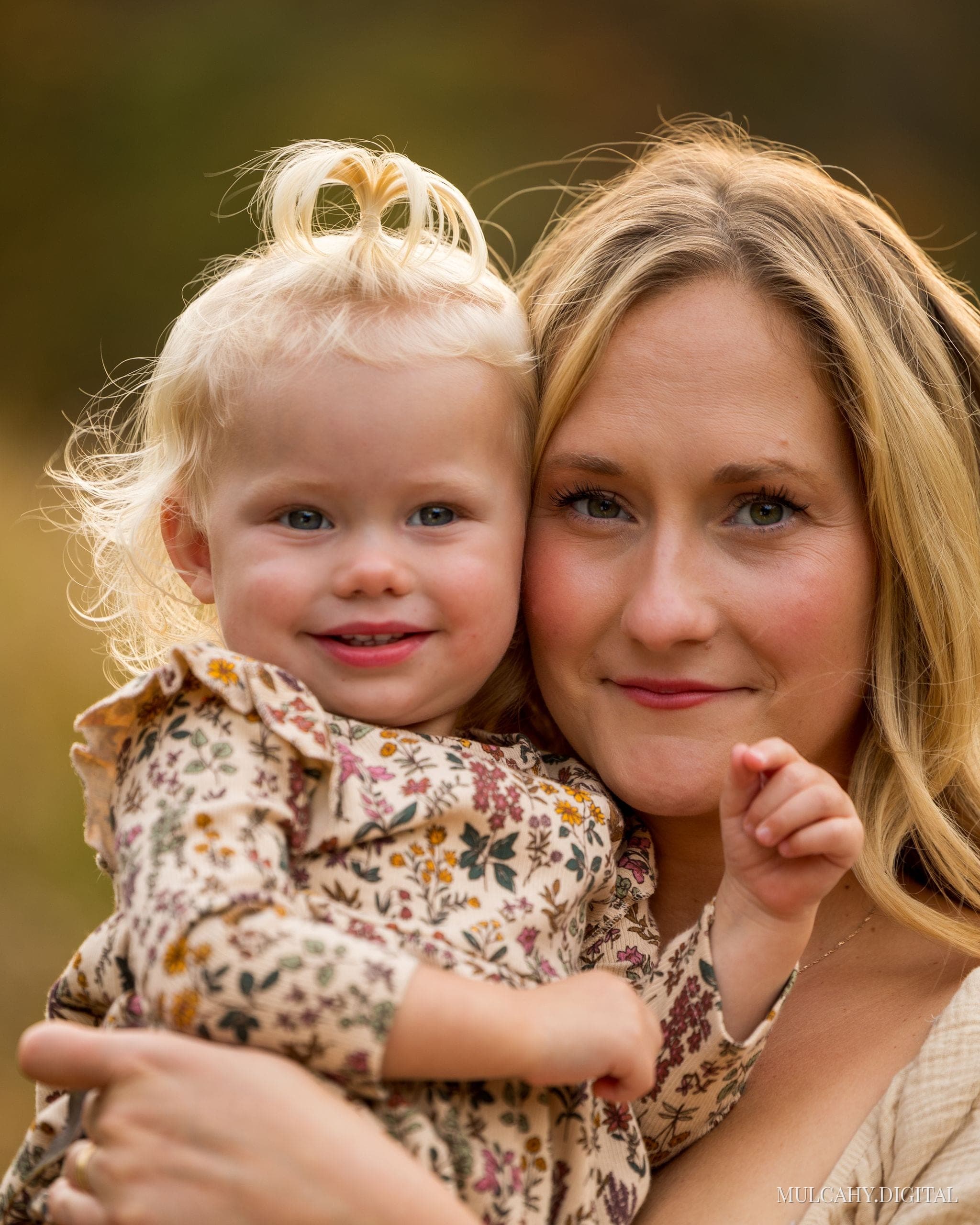 Blond mother cheek to cheek with her toddler daughter in a floral dress, warm autumn light