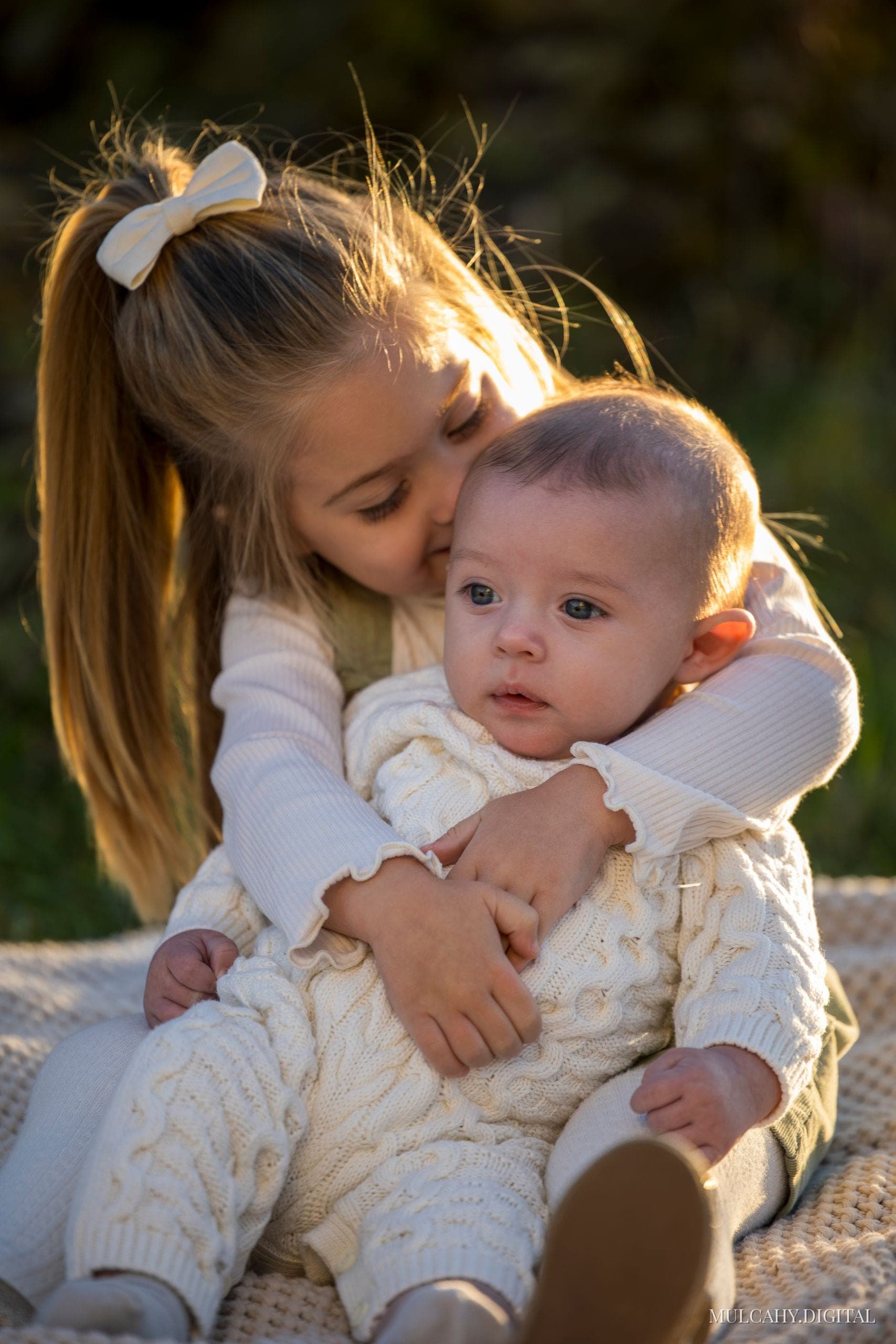 Young girl with a hair bow kissing her baby sibling on a knit blanket at golden hour