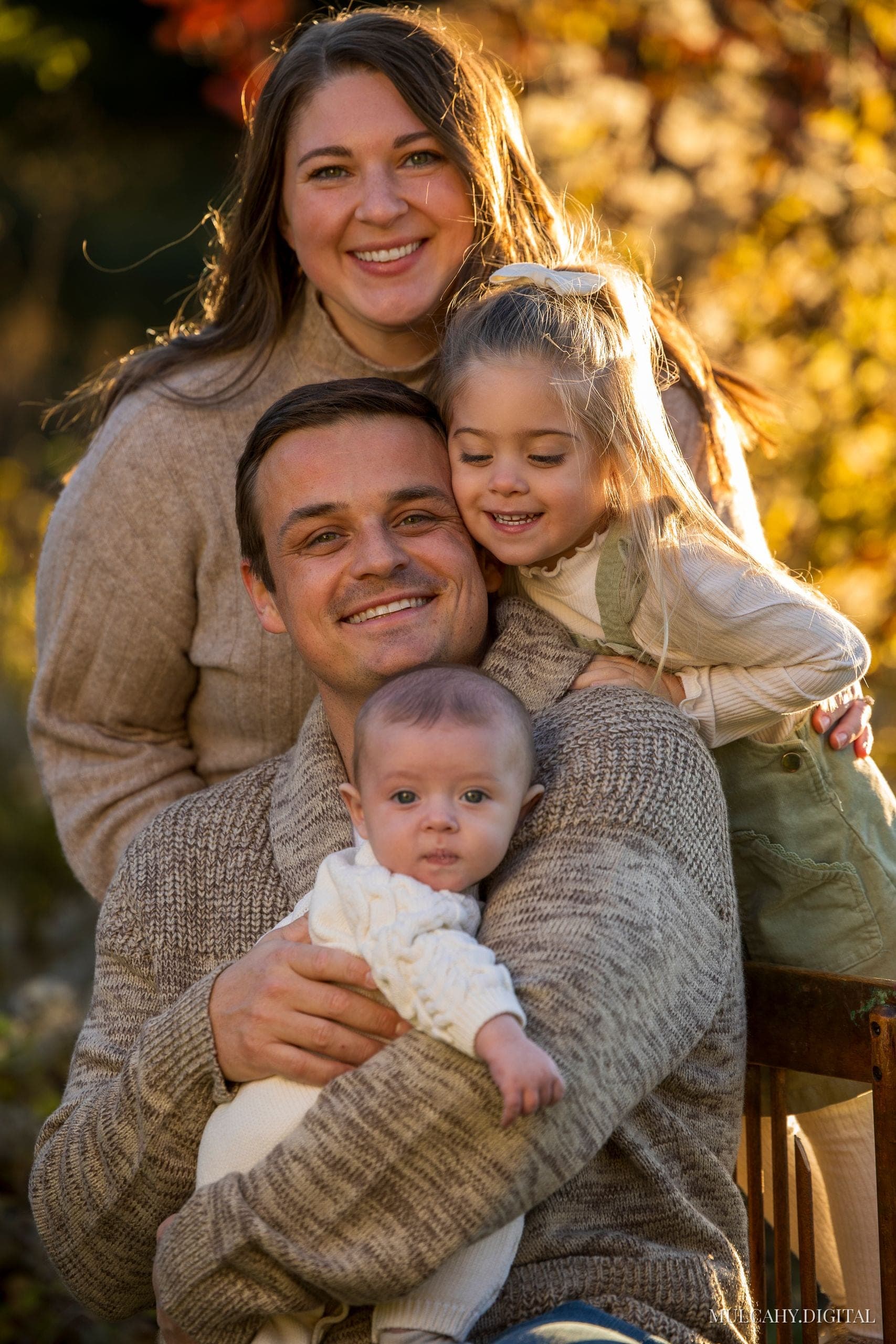 Family of four embracing among autumn foliage, father holding the baby with their young daughter and mother close beside