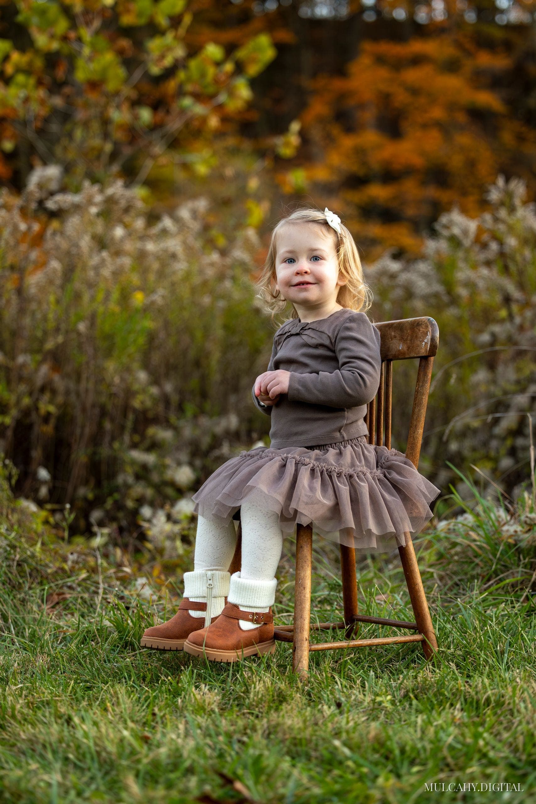 Toddler girl in a tulle dress and boots perched on a wooden chair in an autumn field