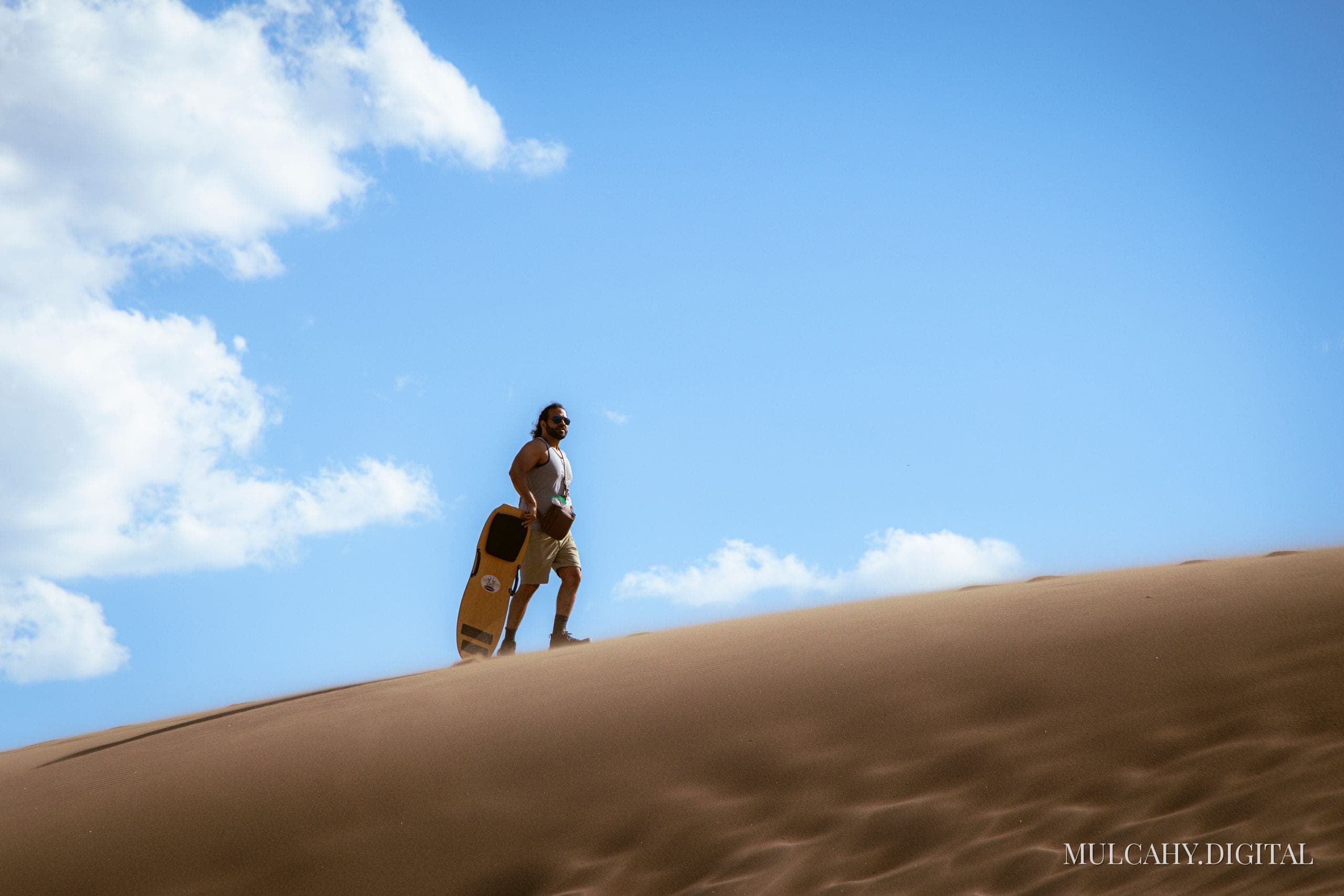 Sandboarder standing atop a sand dune holding a board against a deep blue sky