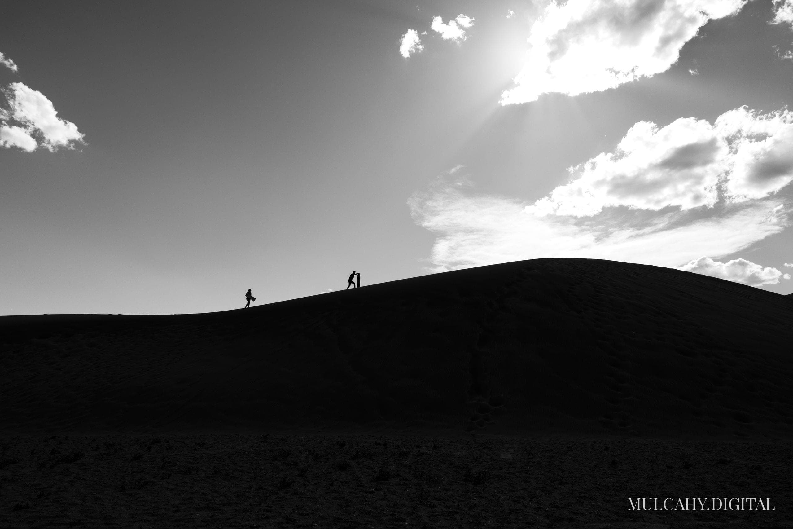 Two people silhouetted on a sand dune ridge against a bright sunburst sky, in black and white